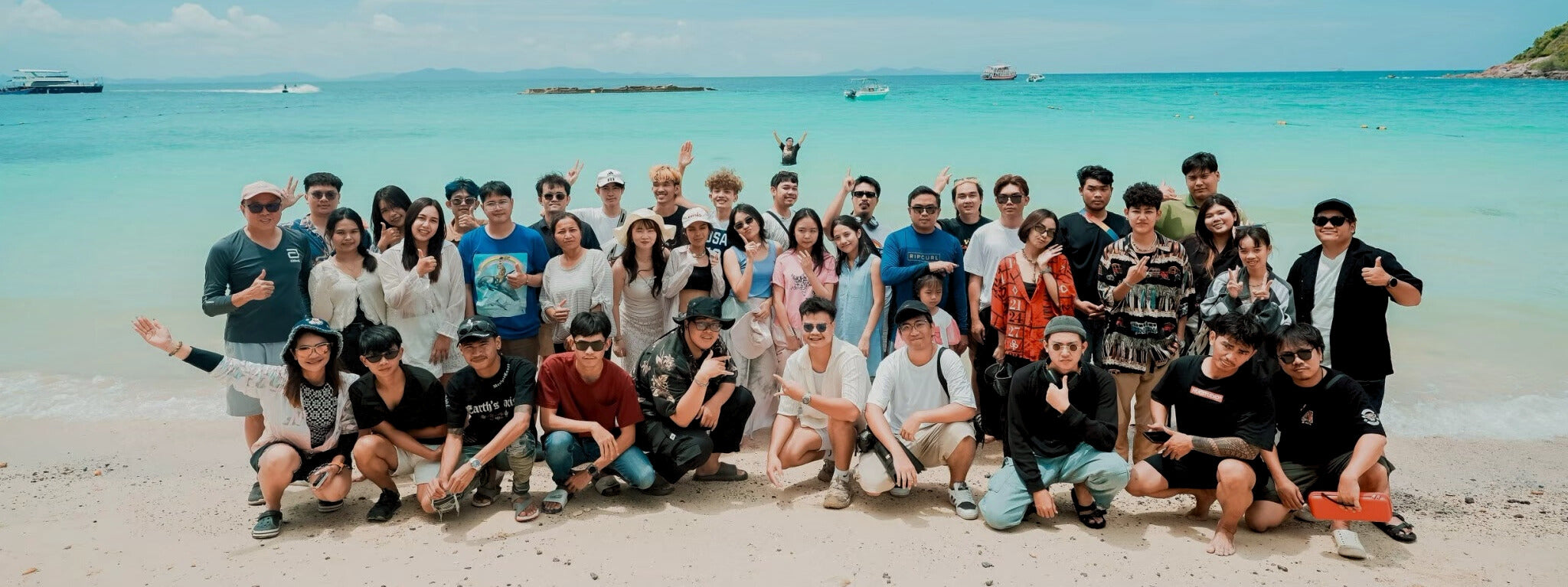 HUGCUSTOM posing for a photo on a beach with clear blue water and a boat in the distance.